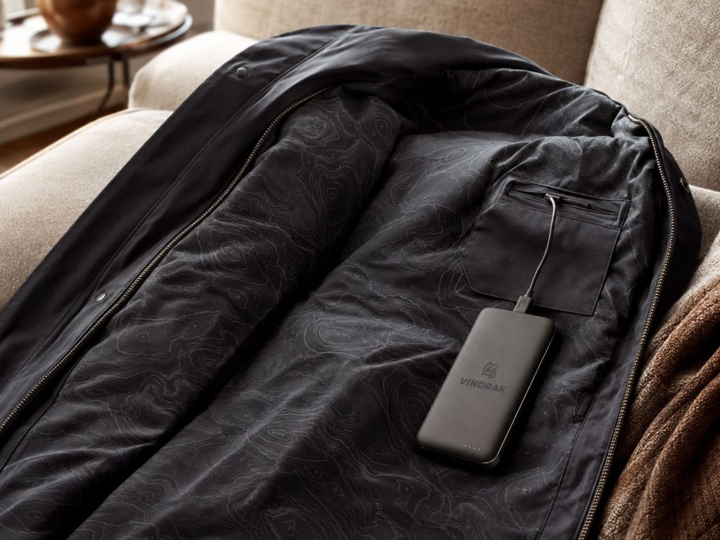 Åpen svart varmejakke på en sofa, med batteriet koblet til i innerlommen